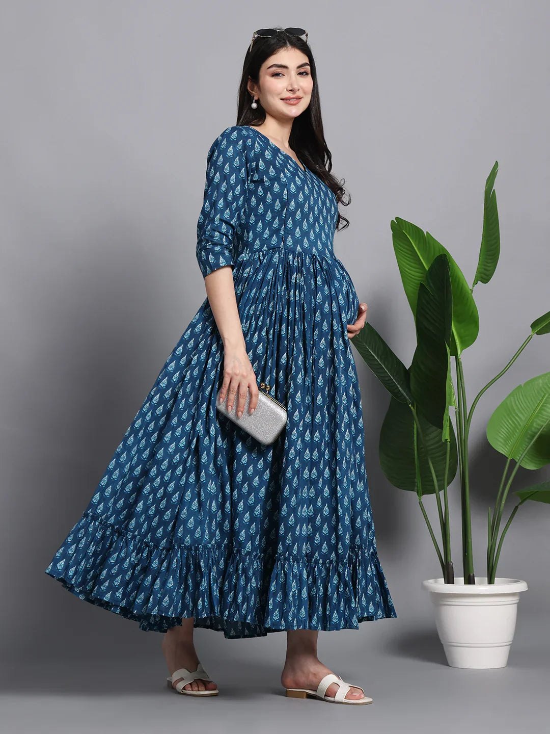 Pregnant woman in blue printed maternity dress, holding clutch, standing by potted plant
